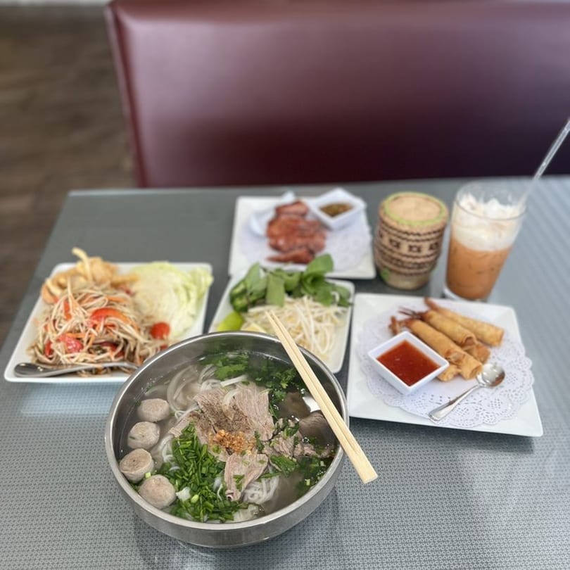 A table spread with Thai dishes including noodle soup, papaya salad, spring rolls, and Thai iced tea.
